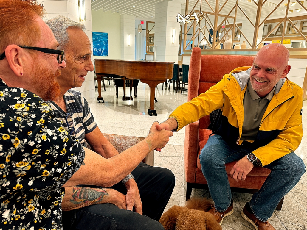 Two men seated on couch shaking hands with seated leader in raincoat