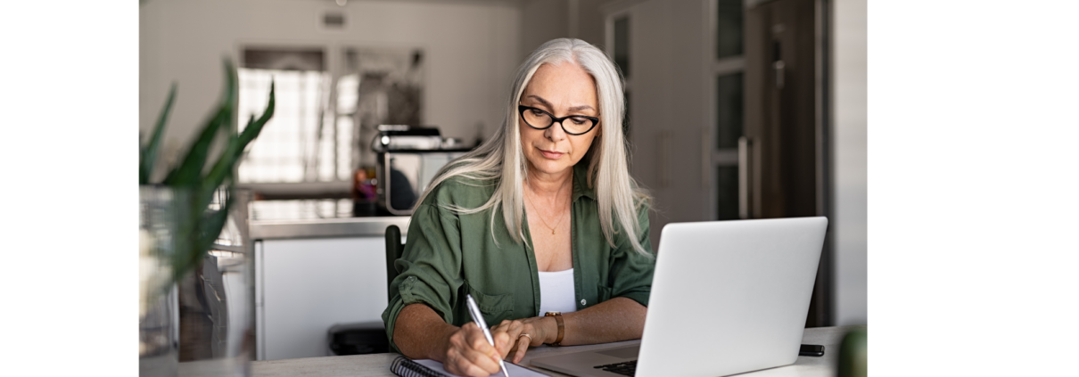 older woman with long hair taking notes sitting at a desk in front of a laptop