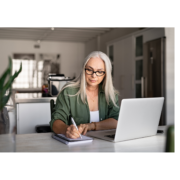 older woman with long hair taking notes sitting at a desk in front of a laptop